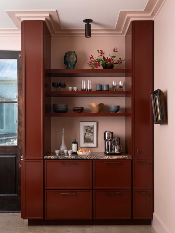 A deep red built-in bar with open shelving, a marble counter and glassware, against pale pink walls.