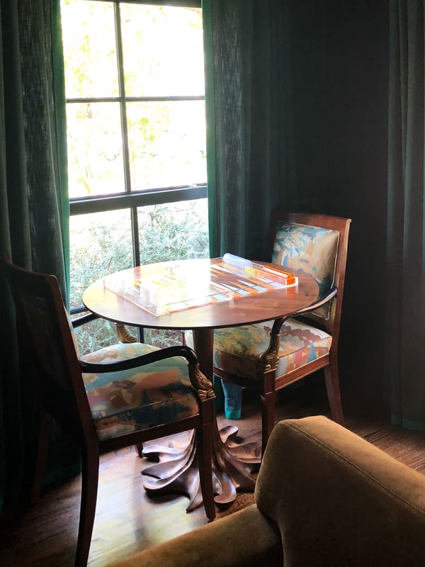A round game table and two upholstered chairs by a window hung with deep green curtains.