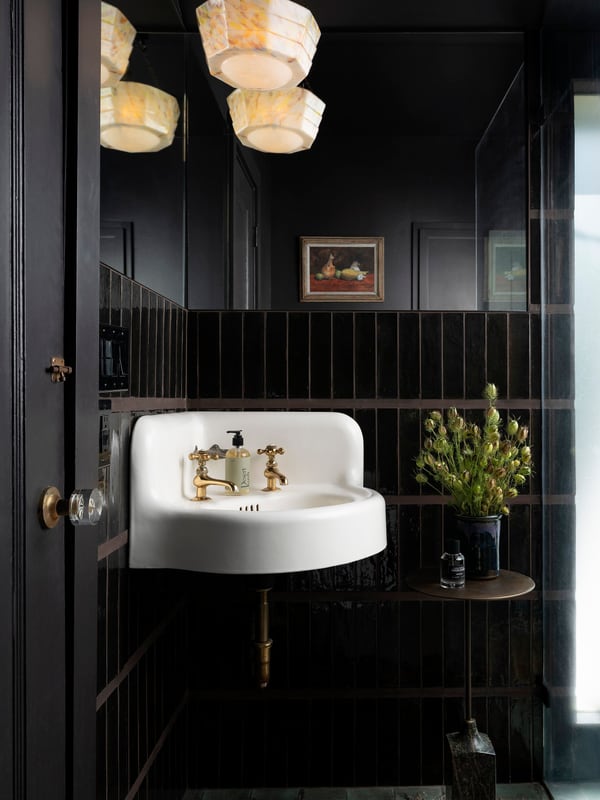 A powder room lined in glossy black tile, with a wall-hung sink, brass taps and a small still life above it.