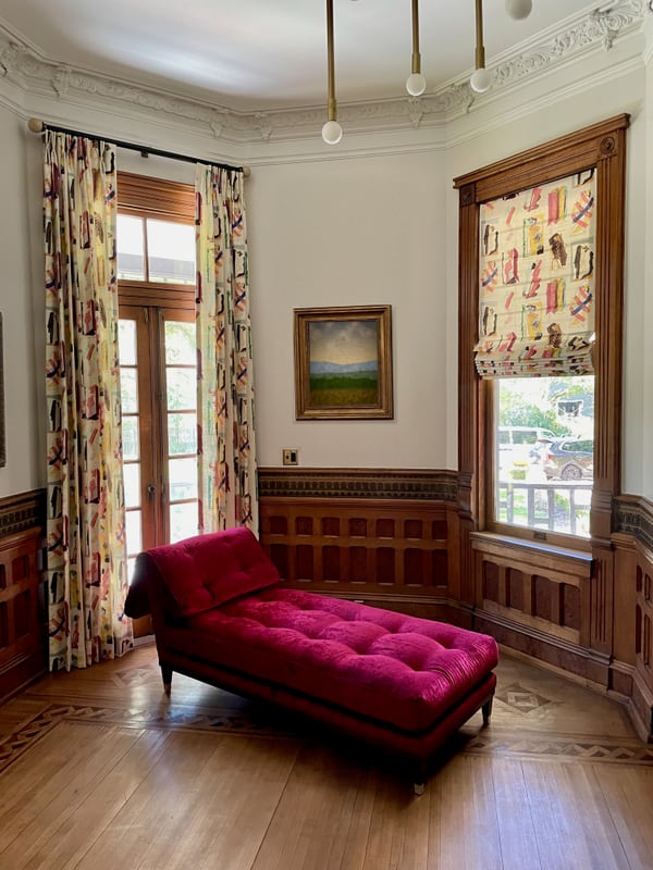 A raspberry velvet chaise in a wood-panelled room with patterned curtains and framed portraits.
